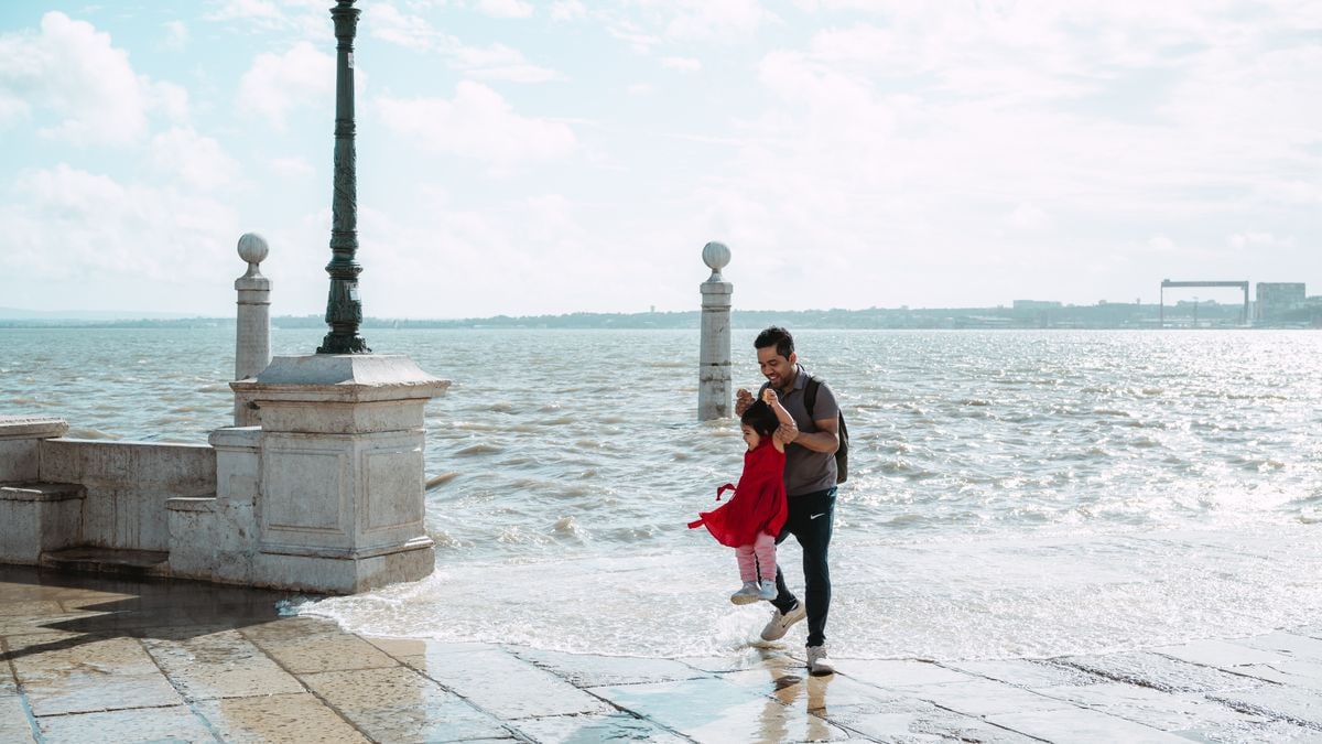 A father joyfully swings his daughter by the sea on a bright day, capturing a playful moment.